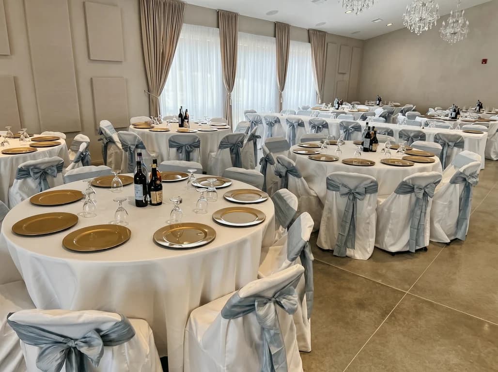 Wide view of round banquet tables with gold chargers and dusty blue chair sashes set for a reception