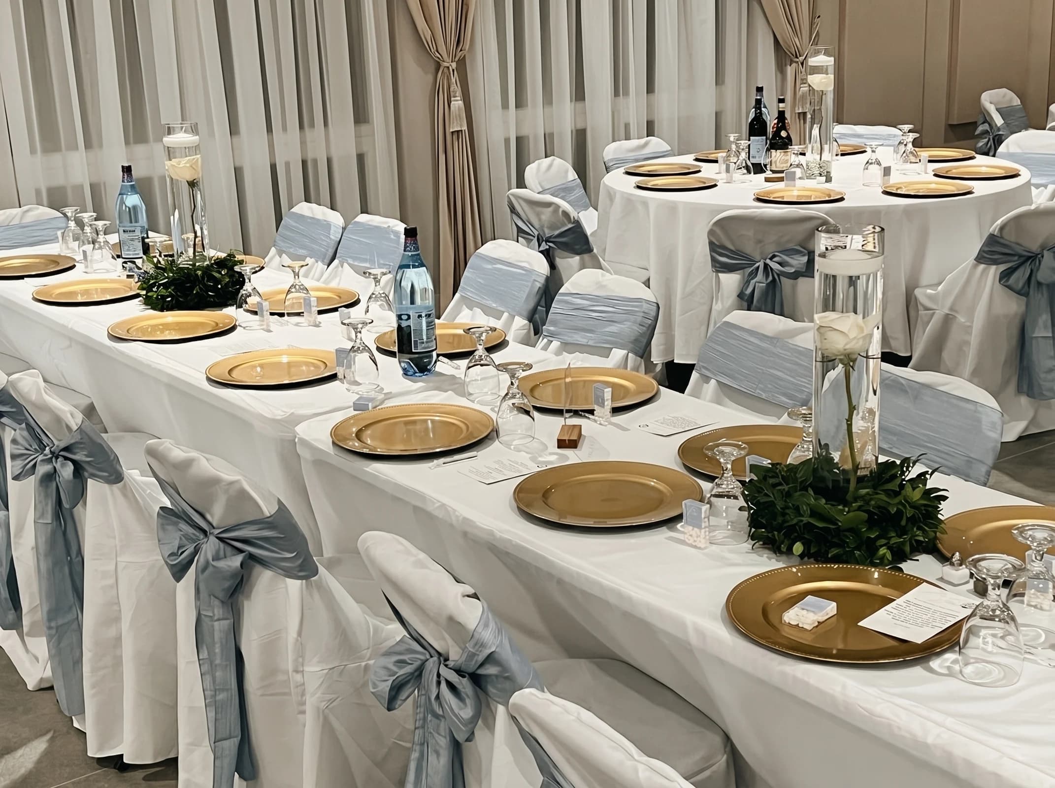 Long banquet table with gold chargers, greenery centerpieces and dusty blue chair sashes at Fellowship Event Hall